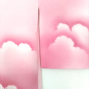 May include: Close-up of a pink fabric featuring a cloud design. White clouds are set against a gradient of pink shades. The fabric is folded, showing a white edge.