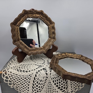 May include: Two octagonal mirrors featuring ornate, textured gold-toned frames with scalloped edges. One mirror is displayed upright on a dark brown stand, while the second mirror lies flat. Both pieces exhibit a reflective glass surface.