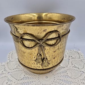 May include: A vintage brass planter with a textured surface and rope detailing. The planter features a decorative rope bow with tassels. The brass has a warm, aged patina, adding to its antique charm. This decorative piece is suitable for indoor or outdoor use.