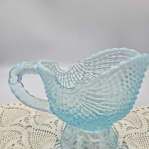 May include: A light blue glass pitcher with a textured, shell-like design. The pitcher has a handle and a wide spout. The base is a pedestal with a floral pattern. The pitcher is sitting on a white lace doily.
