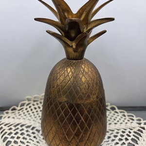 Puede incluir: Un recipiente de latón con forma de piña y tapa extraíble. El cuerpo presenta una superficie texturizada con un patrón de diamantes, y la parte superior está adornada con hojas puntiagudas erguidas. El metal tiene un acabado patinado.