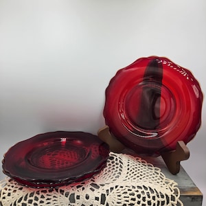 May include: A set of deep red glass plates featuring a scalloped edge and a circular centre design. One plate is displayed upright on a brown stand, while two identical plates are stacked horizontally in front of it, showcasing the translucent glass.
