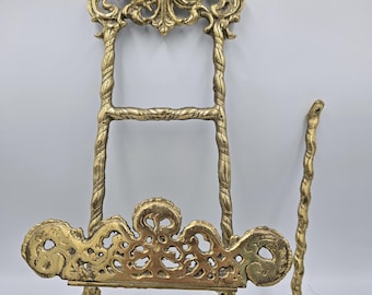 May include: Ornate gold-colored easel with intricate scrollwork and a decorative top featuring birds. The easel has a twisted rope-like frame and a separate support rod. The design is vintage and detailed.