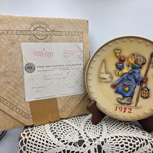 May include: A 1972 commemorative collector plate featuring a relief of a figure in a blue coat holding a horn and lantern, displayed on a brown stand next to a tan mailing envelope from The Bradford Exchange with printed address information.