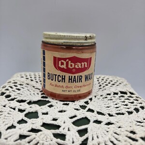 Pode incluir: Um frasco de vidro vintage de cera para cabelo Q'ban Butch Hair Wax com tampa branca de rosca. O rótulo apresenta texto em vermelho e azul sobre fundo bege, indicando Q'ban Butch Hair Wax for Butch, Burr, Crew Haircuts e peso líquido 3 1/4 oz.