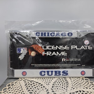 Puede incluir: Un marco de matrícula de los Chicago Cubs cromado en un embalaje de plástico transparente. La barra superior presenta la palabra CHICAGO en azul, mientras que la inferior muestra el logo y nombre de los Cubs. Se ve un guante y una pelota.