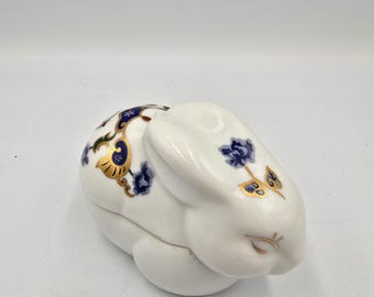 May include: A white ceramic bunny-shaped trinket box. The box features blue floral and gold accents on the ears and back. The bunny is in a seated position with its ears upright. The box is on a white surface.
