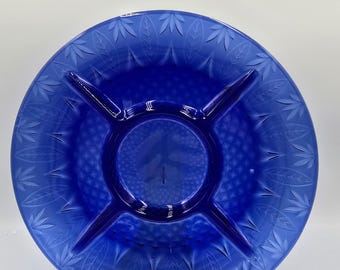 May include: A cobalt blue glass serving dish with a central circular section and four radiating dividers. The outer rim features a leaf pattern, and the center has a textured design. The dish is displayed on a clear stand.