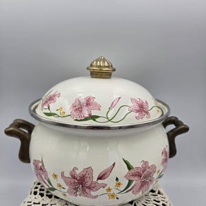 May include: A white enamel pot with a lid, featuring a floral pattern of pink lilies and small yellow flowers. The pot has two dark metal side handles and a matching dark metal knob on the lid. The rim of the pot and lid are finished in silver.