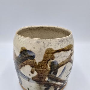 May include: A handmade ceramic tumbler with a speckled off-white glaze. The exterior features abstract brushstrokes in brown and blue. The cylindrical cup has a wide opening and a natural, unglazed base. This unique piece is suitable for beverages.