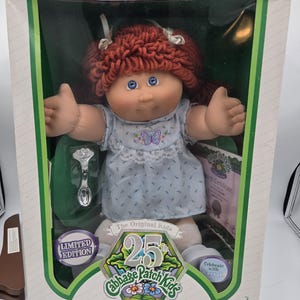 May include: A Cabbage Patch Kids doll with red yarn hair and blue eyes, wearing a light blue dress with a butterfly print. The doll is in a 25th Anniversary limited edition box with a silver spoon, birth certificate, and text reading I'm One of a Kind.