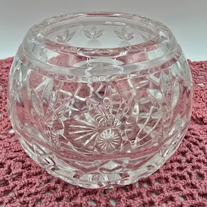 May include: A clear, round crystal bowl with intricate floral and geometric cut patterns. The bowl has a wide opening and sits on a pink crocheted surface. The crystal reflects light, showcasing the detailed design.