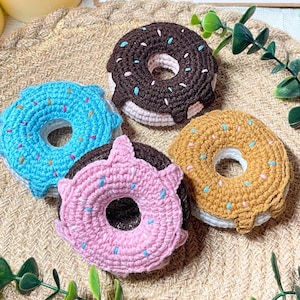 May include: Four colorful, crocheted donut coasters in shades of blue, pink, brown, and tan, each with a white base and sprinkles. The coasters are arranged on a woven mat, with greenery and candles in the background. The text "Grandma Crochet" is visible.