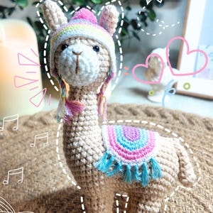 May include: A crocheted llama figurine with a tan body, wearing a colorful hat and saddle blanket. The hat features a pink pom-pom and rainbow stripes. The saddle blanket has blue tassels. The llama is standing on a textured brown surface.