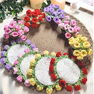 May include: A handmade crochet floral wreath with matching coasters. The wreath is adorned with red, pink, purple, and yellow crocheted flowers. The coasters are white with green and colorful flower accents. The wreath and coasters sit on a brown woven mat.