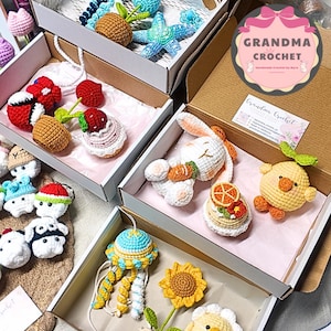 May include: A collection of handmade crochet items, including a blue jellyfish, a yellow sunflower, and a white bunny with a carrot, displayed in open cardboard boxes. Other items include a blue starfish, a red strawberry, and a yellow chick.