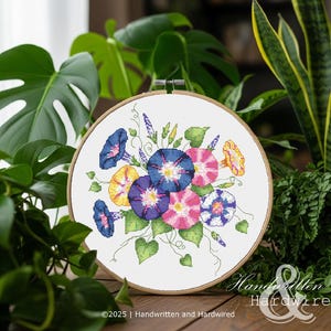 Könnte beinhalten: Ein Kreuzstich-Stickrahmen mit einem Blumendesign aus Prunkwinden in Blau-, Rosa-, Gelb- und Lilatönen. Die Stickerei ist vor einem weißen Hintergrund und von einem Holzreifen umrahmt. Der Text "Handwritten & Hardwired" ist sichtbar.