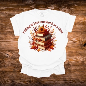 May include: A white t-shirt featuring a graphic of a stack of four books topped with a sunflower and small red apples. The design is surrounded by autumn leaves and the text "Falling in love one book at a time" in a brown, curved font.