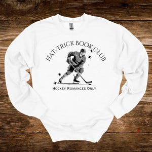 Pode incluir: Camisola branca com uma ilustração a preto e branco de um jogador de hóquei. O texto "HAT-TRICK BOOK CLUB" está arqueado acima da imagem, com "HOCKEY ROMANCES ONLY" abaixo. O design inclui estrelas.
