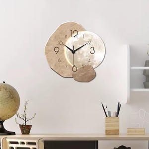 May include: A contemporary wall clock featuring a moon-like design. The clock face is a white circle with black hands and numerals, set against beige, organic shapes. The clock is mounted on a white wall, adding a modern touch to any room.