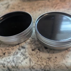 Puede incluir: Dos pequeñas latas redondas plateadas, una abierta y llena de una sustancia negra oscura, la otra cerrada. Las latas están sobre una superficie clara y moteada.