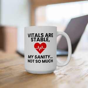 May include: White ceramic mug with the text "VITALS ARE STABLE, MY SANITY... NOT SO MUCH" in black and red. The mug features a red heart with a white heartbeat line. The mug is on a wooden surface.