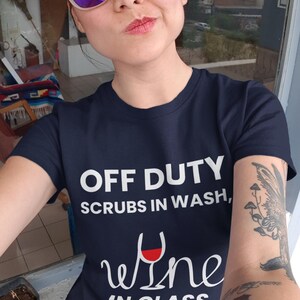 May include: Navy blue t-shirt with the text "OFF DUTY, SCRUBS IN WASH, wine IN GLASS" in white. The shirt features a wine glass graphic with red wine. The person is wearing purple sunglasses.
