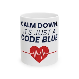 May include: White ceramic mug with the text "CALM DOWN, IT'S JUST A CODE BLUE" in navy blue. A red heart with a white EKG line is centered below the text. A red line extends from the heart to both sides of the mug.