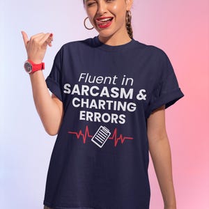 May include: Navy blue t-shirt with the text "Fluent in Sarcasm & Charting Errors" in white. A red heartbeat line graphic with a chart icon is below the text. The person is wearing a red watch.