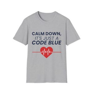 May include: Gray t-shirt with the text "CALM DOWN, IT'S JUST A CODE BLUE" in navy blue. A red heart with a heartbeat graphic is centered below the text, with a red line extending from the heart.