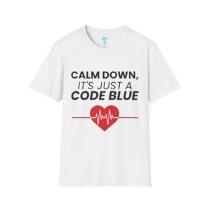 May include: White t-shirt with the text "CALM DOWN, IT'S JUST A CODE BLUE" in black. Below the text is a red heart with a heartbeat line. The shirt is a crew neck style.
