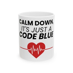 May include: White ceramic mug with the text "CALM DOWN, IT'S JUST A CODE BLUE" in black. A red heart with a white EKG line is centered below the text. The mug is a standard size and shape.