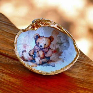 May include: A decorative seashell with a painted image of a teddy bear wearing a bow tie. The shell has a gold-colored rim and is set on a wooden surface. The teddy bear is brown and white with a pink bow tie.