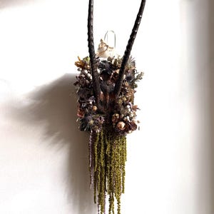 May include: A wall-mounted display featuring two long, dark, curved animal horns attached to a rectangular base. The base is adorned with a dense arrangement of dried flowers, foliage, and long, cascading green amaranth stems hanging downward.