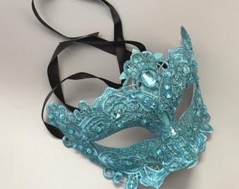 May include: A teal lace masquerade mask with sparkling accents and a black ribbon tie. The mask features intricate floral patterns and is embellished with clear, iridescent gemstones. This decorative mask is suitable for costume parties and formal events.
