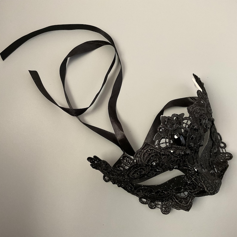 May include: Black lace masquerade mask with a decorative design. The mask features a central jewel and is attached to black satin ribbons for tying. The mask is designed for a formal event or costume.