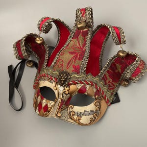 May include: A decorative Venetian-style jester mask with red velvet and gold brocade fabric. The mask features a cracked ivory finish, musical notes, and red diamond accents. Gold bells hang from the jester's hat.