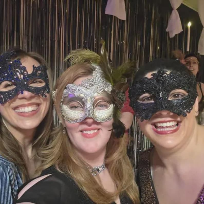 May include: Three smiling people wearing decorative masquerade masks. The masks are black, silver, and gold, with intricate lace and feather details. The background features a festive setting with hanging decorations.