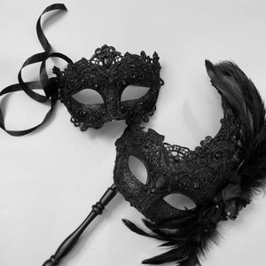 May include: Two black masquerade masks. One is a full face mask with a ribbon tie. The other is a half mask with a handle and black feathers. Both masks have intricate lace and glitter details.