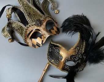 May include: Two ornate Venetian masks. One is a jester mask with gold, black, and cream colors, featuring bells and a harlequin pattern. The other is a gold and black mask with a handle and black feathers.
