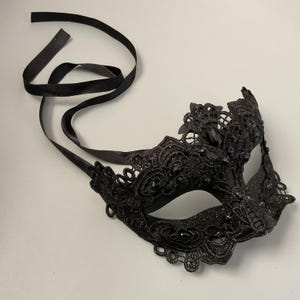 May include: Black lace masquerade mask with a delicate floral design and attached black satin ribbons for tying. The mask features intricate detailing and sparkling accents, perfect for a formal event or costume party.