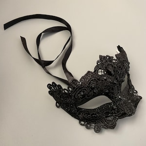 May include: Black lace masquerade mask with a decorative floral design and black satin ribbon ties. The mask features intricate detailing and is suitable for costume parties or formal events.
