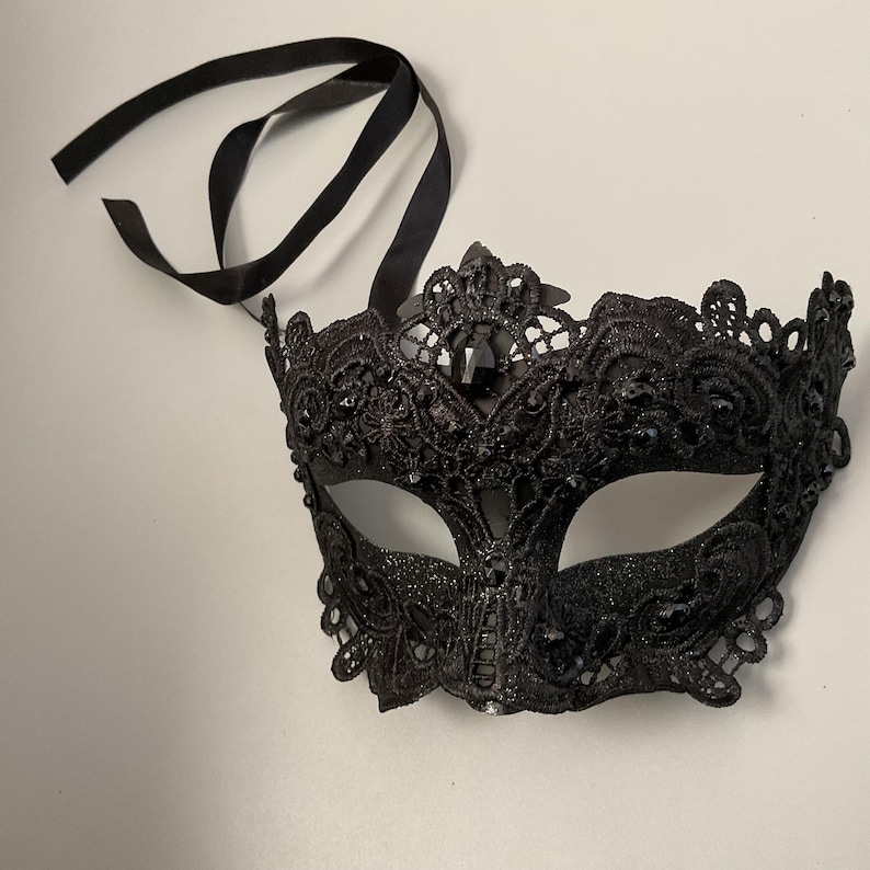 May include: A black masquerade mask with intricate lace detailing and sparkling accents. The mask features a decorative crown design and is attached to black satin ribbons for securing. A glamorous accessory for parties and events.
