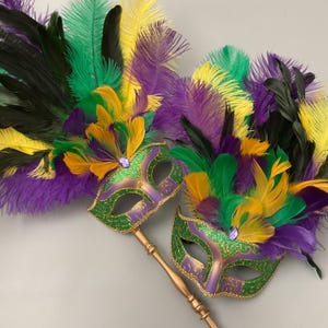 May include: Two Mardi Gras masks with purple, green, yellow, and black feathers. The masks are purple with green and gold glitter accents and a jewel. Each mask has a gold-coloured handle.