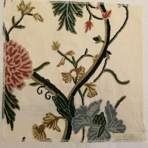 May include: A square piece of off-white fabric featuring an embroidered floral pattern with large pink and blue blossoms, dark green leaves, and thin brown stems. A small rectangular label is attached to the bottom right corner of the textile.
