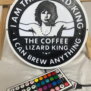 May include: A round, black and white sign with the text "I AM THE LIZARD KING I CAN BREW ANYTHING" and an image of a person. A remote control with colorful buttons is in the foreground.