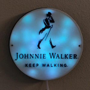 May include: Round, illuminated sign featuring the Johnnie Walker logo: a black silhouette of a man walking with a cane. The sign has a white background with blue lighting and the text "JOHNNIE WALKER. KEEP WALKING."