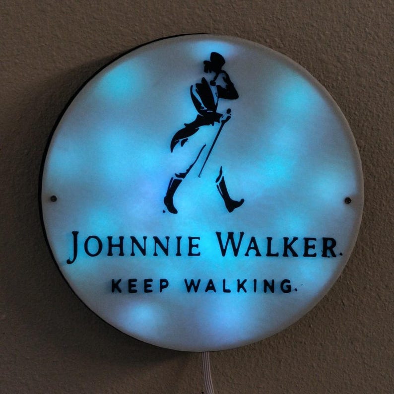 May include: Round, illuminated sign featuring the Johnnie Walker logo: a black silhouette of a man walking with a cane. The sign has a white background with blue lighting and the text "JOHNNIE WALKER. KEEP WALKING."