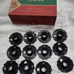 May include: A set of twelve black plastic sewing machine cams arranged in three rows, accompanied by a rectangular cardboard box labelled SINGER Special Discs for classes 600 and 603 machines, featuring green and red panels with white text.