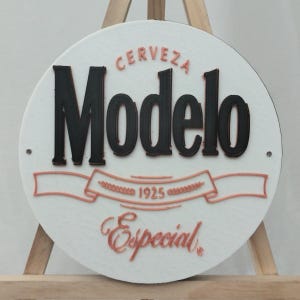 May include: A round white sign with the word "Modelo" in large black letters. The words "CERVEZA" and "Especial" are in a copper color, along with the year "1925" and a banner design. The sign is mounted on a wooden easel.